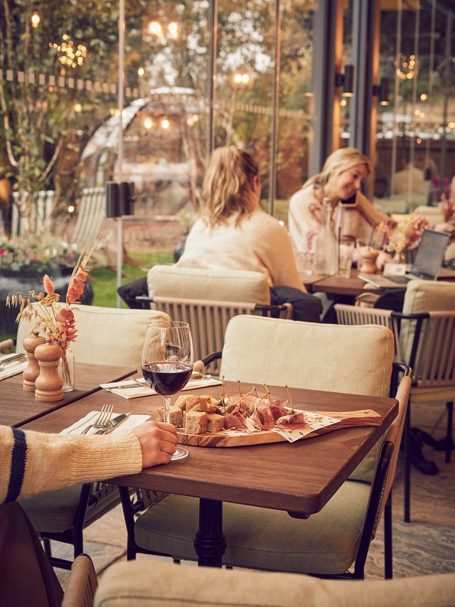 Hand holds a glass of wine in front of a sharing platter, two women sit in the background