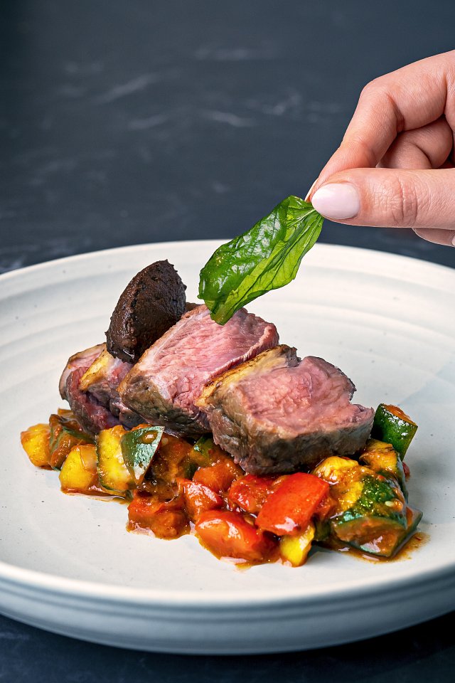 Lamb rump on a bed of vegetables, with a lady's hand adding a basil leaf