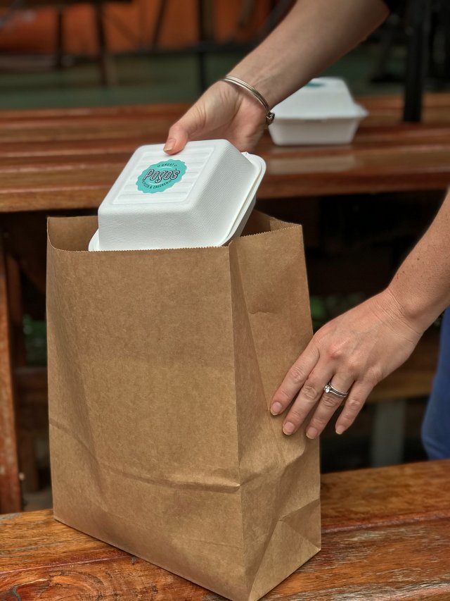 Hand placing a polystyrene takeaway box, saying Pogo's – great pizza and chicken – into a brown paper bag