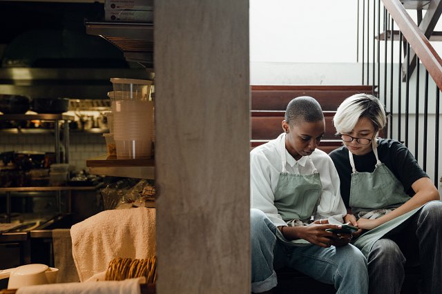 Two waitresses sit on the stairs behind a kitchen, looking at a phone